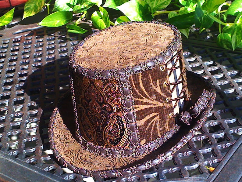 May include: A brown and gold top hat with a patterned design and a brown leather band. The hat has a lace-up detail on the side.