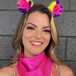 May include: A woman wearing a pink bandana with rainbow fringe and fuzzy pink and yellow cat ears.