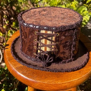 May include: A brown top hat with a patterned leather top, brown velvet sides, and a brown and beige striped ribbon with gold eyelets and laces.