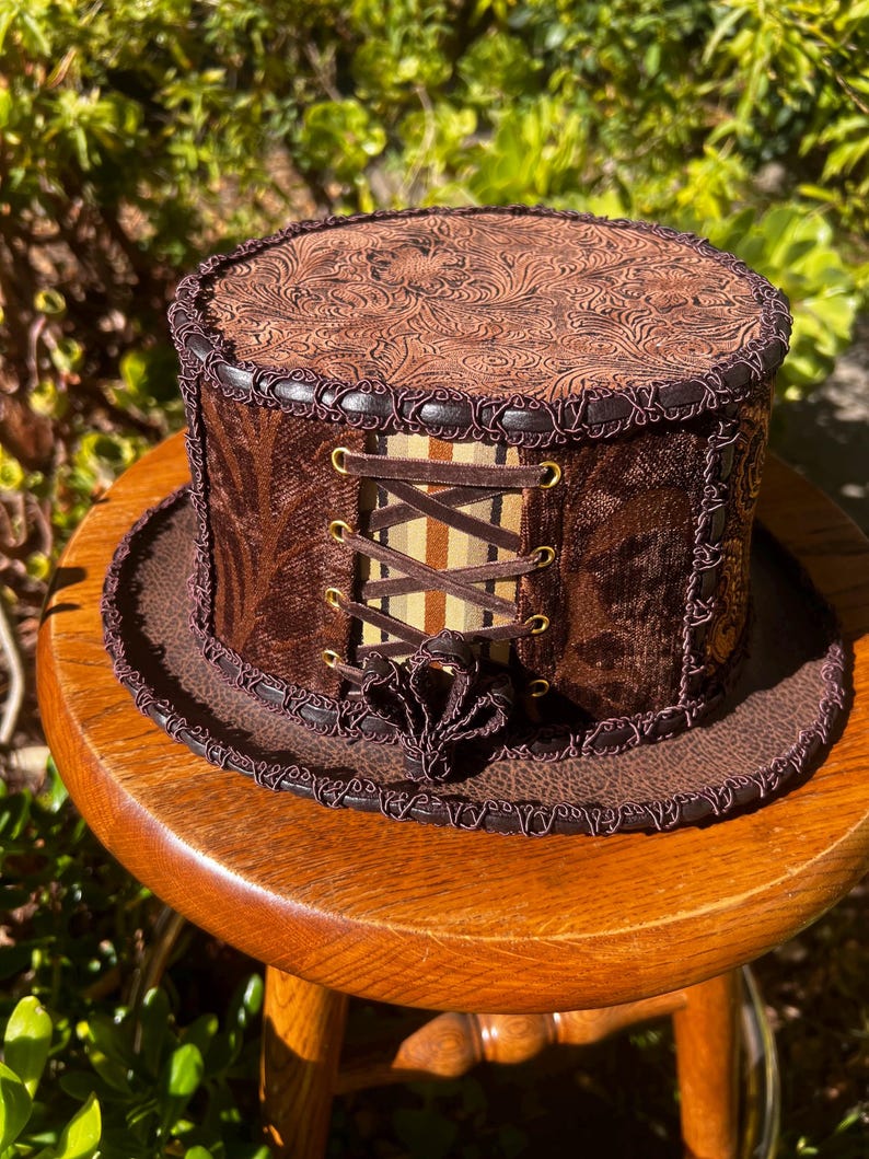 May include: A brown top hat with a patterned leather top, brown velvet sides, and a brown and beige striped ribbon with gold eyelets and laces.