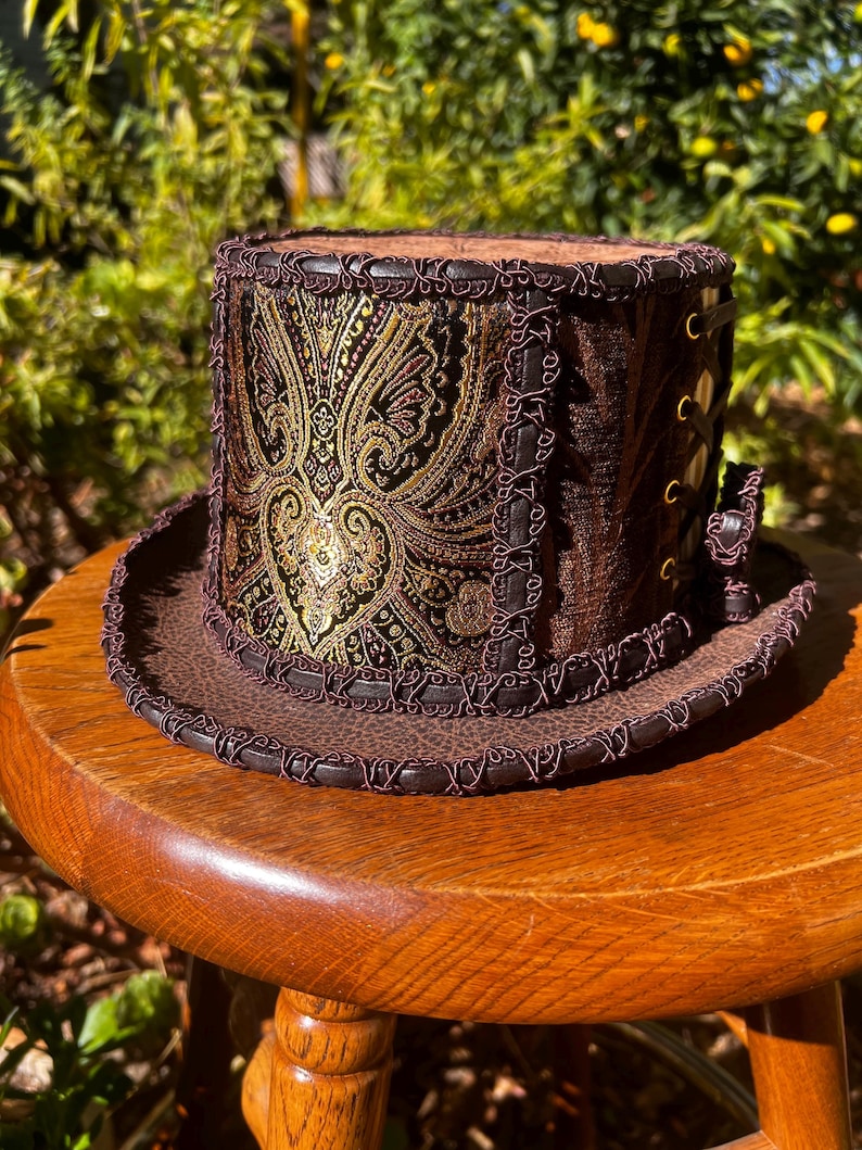May include: A brown top hat with a gold and black paisley pattern on the crown. The hat has a brown leather brim with a decorative stitching detail.