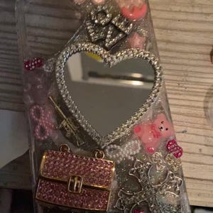 May include: Clear phone case decorated with various charms, including a heart-shaped mirror, a pink purse, a cross, and the word "DIOR". The case is embellished with rhinestones and pink accents, creating a glamorous aesthetic.