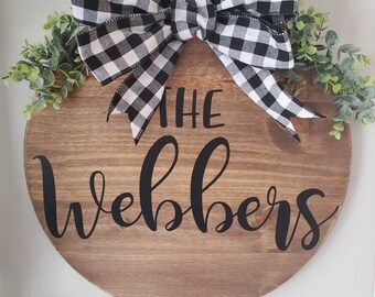 Può includere: Un cartello rotondo in legno con il testo nero "THE Webbers" al centro. Un grande fiocco a quadri bianchi e neri è fissato sulla parte superiore, affiancato da foglie di eucalipto artificiali verdi. Una corda di iuta funge da gancio.