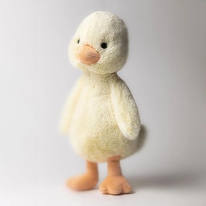 May include: A plush, soft, yellow duck toy with a light orange beak and feet. The duck has small black eyes and a fluffy texture. The toy is standing upright against a white background, showcasing its full form.