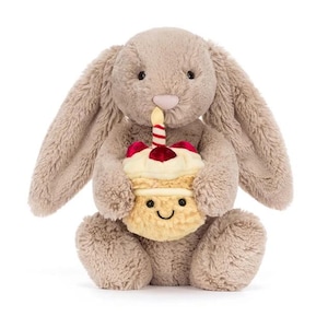 May include: A plush, light brown bunny holding a cupcake with a lit candle. The cupcake has a smiling face, red cherry topping, and a striped candle. The bunny has long ears and a pink nose.