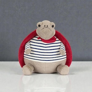 May include: A plush turtle toy with a tan body, red shell, and a striped blue and white shirt. The turtle has a red scarf around its neck and is sitting on a white surface against a gray background.