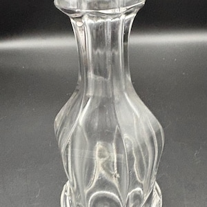 May include: A clear glass vase with a hexagonal top and a rounded, textured body. The vase has a flared base and a narrow neck. The glass is transparent, allowing light to pass through. The vase is likely for decorative purposes.
