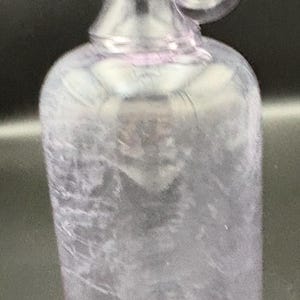 May include: A clear, lavender-tinted glass bottle with a rounded body and a handle. The bottle has a narrow neck and a circular handle attached near the top. The glass has a slightly textured appearance.
