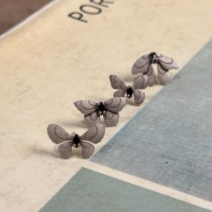 Op de afbeelding: Vier grijze vlindervormige oorbellen met zwarte centra zijn in een rij gerangschikt. De oorbellen liggen op een lichtgekleurd oppervlak met het woord "PORCELAIN" zichtbaar. De vlinders hebben gedetailleerde vleugelpatronen.