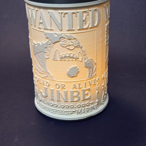 May include: Cylindrical lamp with a textured surface, featuring the text "WANTED" and "JINBE" in a stylized font. The lamp has a light-coloured design with a dark top and base, and a light source at the top.