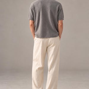 K&ouml;nnte beinhalten: Ein kurz&auml;rmeliges graues Strickshirt mit Rundhalsausschnitt, kombiniert mit einer locker sitzenden cremefarbenen Hose und dunklen Schuhen. Das Shirt hat einen strukturierten Strickstoff, w&auml;hrend die Hose einen geraden Schnitt aufweist.