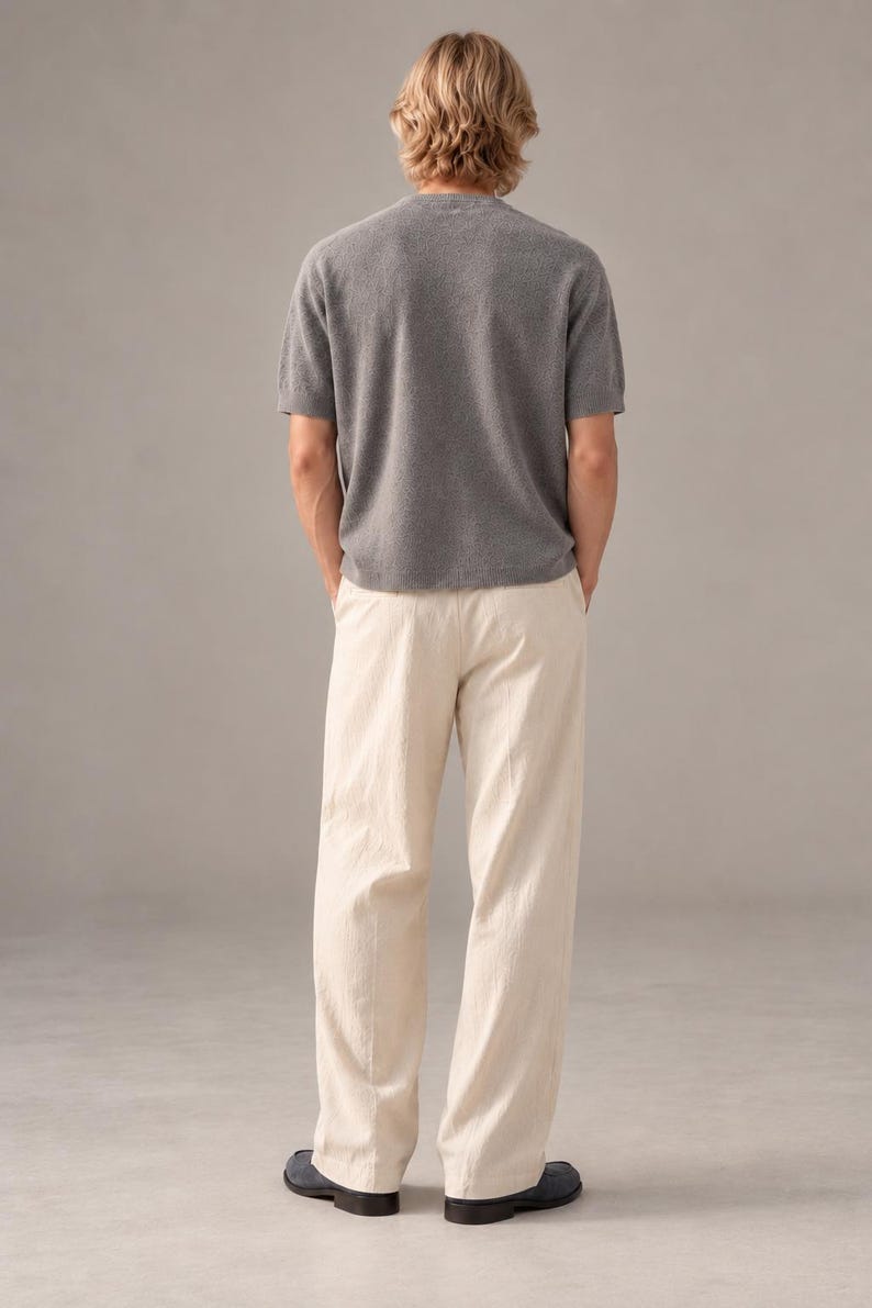 K&ouml;nnte beinhalten: Ein kurz&auml;rmeliges graues Strickshirt mit Rundhalsausschnitt, kombiniert mit einer locker sitzenden cremefarbenen Hose und dunklen Schuhen. Das Shirt hat einen strukturierten Strickstoff, w&auml;hrend die Hose einen geraden Schnitt aufweist.