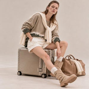 May include: An olive green tie-dye long-sleeve shirt, white shorts, and a cream-colored sweater draped over the shoulders. The outfit includes beige mid-calf boots with buckles, a metallic silver rolling suitcase, and a tan leather duffel bag.