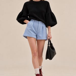May include: A black ribbed knit sweater with voluminous sleeves, light blue tailored shorts with a cuffed hem, white socks, dark red lace-up boots, and a black handbag. The outfit features a round neckline and a structured waistband.