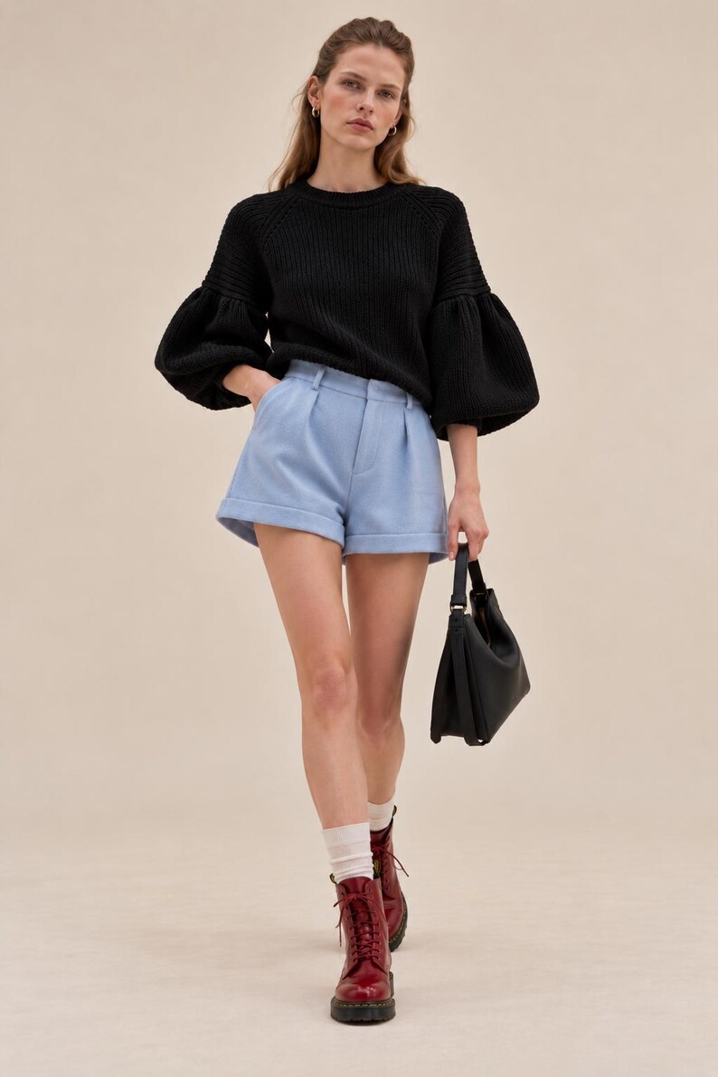 May include: A black ribbed knit sweater with voluminous sleeves, light blue tailored shorts with a cuffed hem, white socks, dark red lace-up boots, and a black handbag. The outfit features a round neckline and a structured waistband.