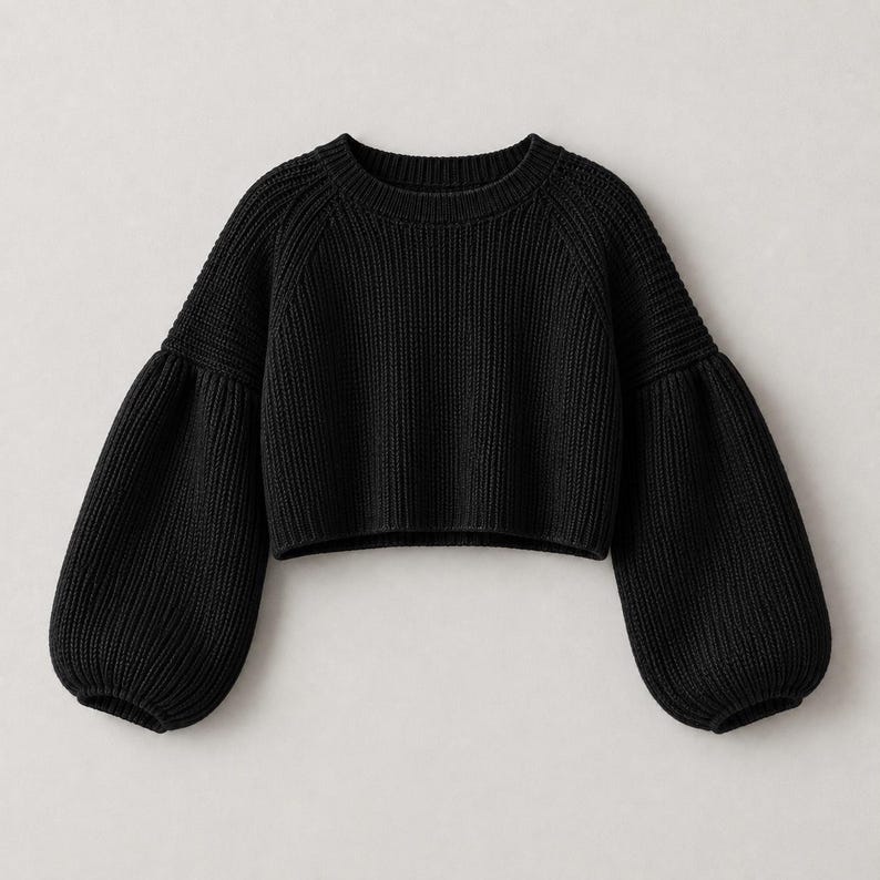 May include: A solid black cropped sweater featuring a ribbed knit texture, a round neckline, and voluminous raglan sleeves that taper into fitted ribbed cuffs. The garment has a straight hemline and a consistent knit pattern throughout.