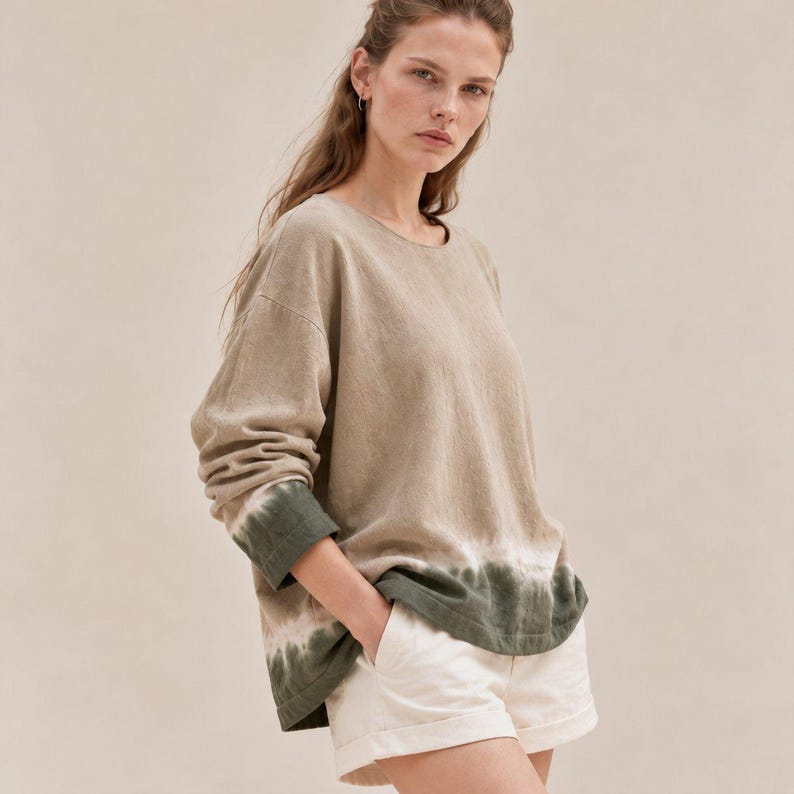 May include: A long-sleeve beige pullover shirt featuring a round neckline and a dark olive green tie-dye pattern along the hem and cuffs, paired with off-white shorts that have a cuffed hem and a visible button closure at the waist.