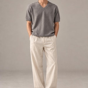 May include: A short-sleeved gray V-neck sweater with a textured knit finish paired with off-white wide-leg trousers featuring a central vertical seam detail and dark blue suede loafers with a classic strap across the top of the foot.