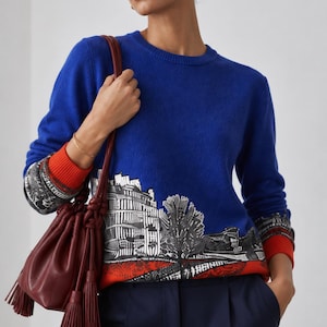 Puede incluir: Un su&eacute;ter de manga larga en color azul real con un estampado gr&aacute;fico en blanco y negro de un paisaje urbano en el dobladillo, acentuado con pu&ntilde;os acanalados de color naranja brillante y una banda naranja a juego en el borde inferior, junto a un bolso de cuero rojo oscuro.