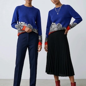 Puede incluir: Un su&eacute;ter azul de manga larga con un gr&aacute;fico de paisaje urbano en blanco y negro en la parte inferior del torso y las mangas, terminado con un dobladillo y pu&ntilde;os de color naranja vibrante. El su&eacute;ter se combina con pantalones azul marino o falda.