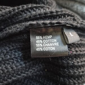 May include: A black knit garment featuring a rectangular fabric tag with the text 55% HEMP, 45% COTTON, 55% CHANVRE, 45% COTON and a separate smaller tag indicating size L. The texture of the black knit fabric is visible around the labels.