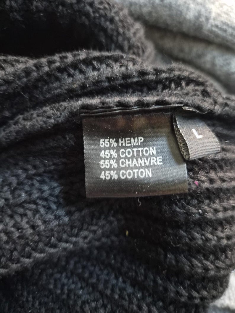 May include: A black knit garment featuring a rectangular fabric tag with the text 55% HEMP, 45% COTTON, 55% CHANVRE, 45% COTON and a separate smaller tag indicating size L. The texture of the black knit fabric is visible around the labels.