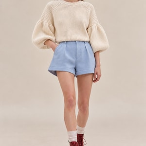 K&ouml;nnte beinhalten: Ein cremefarbener, gerippter Strickpullover mit volumin&ouml;sen langen &Auml;rmeln und Rundhalsausschnitt, kombiniert mit hellblauen, ma&szlig;geschneiderten Shorts mit Faltenfront, umgeschlagenen S&auml;umen und Knopfverschluss, getragen mit wei&szlig;en Socken und dunkelroten Schn&uuml;rstiefeln.