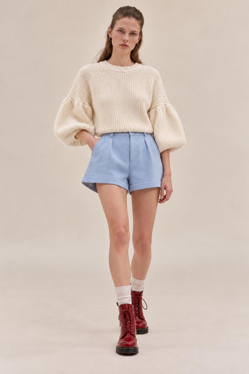 K&ouml;nnte beinhalten: Ein cremefarbener, gerippter Strickpullover mit volumin&ouml;sen langen &Auml;rmeln und Rundhalsausschnitt, kombiniert mit hellblauen, ma&szlig;geschneiderten Shorts mit Faltenfront, umgeschlagenen S&auml;umen und Knopfverschluss, getragen mit wei&szlig;en Socken und dunkelroten Schn&uuml;rstiefeln.