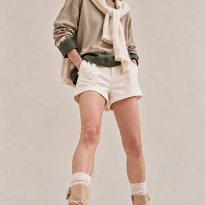 May include: A long-sleeve olive and beige tie-dye sweatshirt with a white sweater draped over the shoulders, paired with white cuffed shorts, white ribbed socks, and tan suede mid-calf boots with side buckles and thick lug soles.