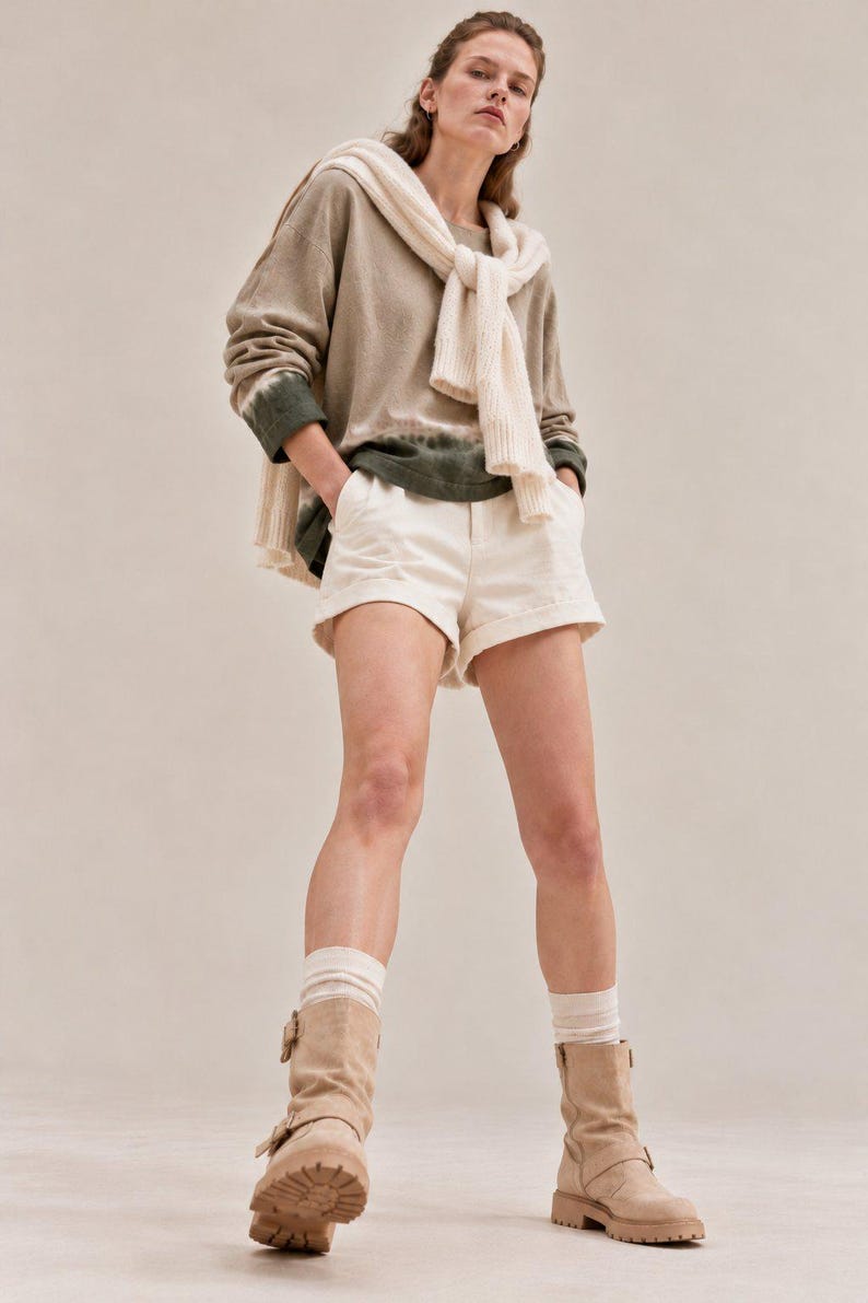 May include: A long-sleeve olive and beige tie-dye sweatshirt with a white sweater draped over the shoulders, paired with white cuffed shorts, white ribbed socks, and tan suede mid-calf boots with side buckles and thick lug soles.