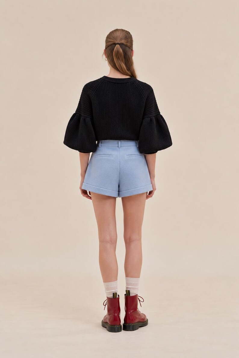 May include: A black long-sleeved sweater with ribbed texture and voluminous bell sleeves paired with light blue tailored shorts featuring a cuffed hem. The outfit is completed with sheer white socks and dark red lace-up ankle boots.