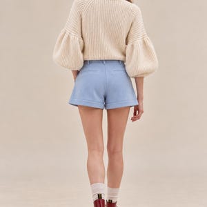 K&ouml;nnte beinhalten: Ein cremefarbener Rippstrickpullover mit volumin&ouml;sen langen &Auml;rmeln kombiniert mit hellblauen, ma&szlig;geschneiderten Shorts mit umgeschlagenem Saum, getragen mit transparenten wei&szlig;en Socken und dunkelroten Schn&uuml;rstiefeln aus Leder mit schwarzen Sohlen.