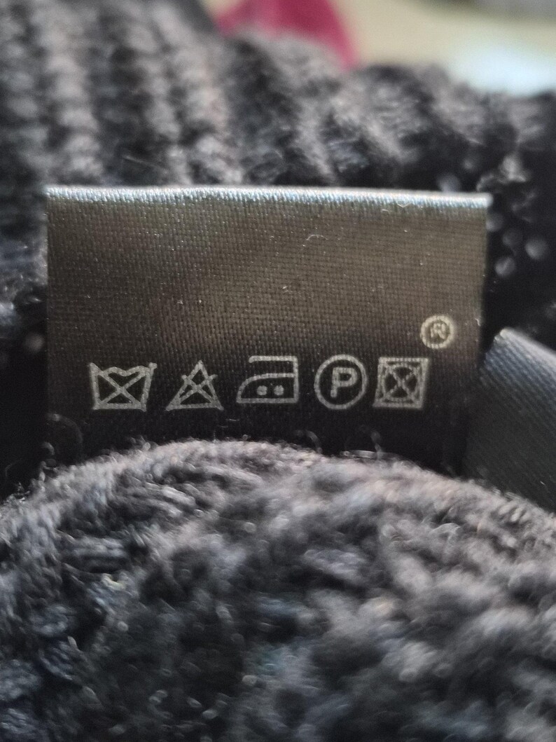 May include: A black fabric garment care label featuring five white laundry symbols: a crossed-out wash tub, a crossed-out triangle, an iron icon, a circle with the letter P, and a crossed-out square with a circle inside.