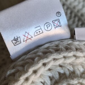 K&ouml;nnte beinhalten: Ein wei&szlig;es Pflegeetikett an einem beigen Strickkleidungsst&uuml;ck. Das Etikett zeigt schwarze Pflegesymbole, darunter ein Waschsymbol f&uuml;r 30 Grad Celsius, ein durchgestrichenes Dreieck, ein B&uuml;geleisensymbol mit zwei Punkten, ein P im Kreis und ein durchgestrichenes Quadrat.