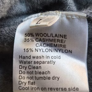K&ouml;nnte beinhalten: Ein wei&szlig;es Textilpflegeetikett an einem grauen Strickstoff. Das Etikett zeigt die Gr&ouml;&szlig;e L und die Materialzusammensetzung von 50 % Wolle, 35 % Kaschmir und 15 % Nylon sowie spezifische Anweisungen zur Handw&auml;sche und Pflege des Kleidungsst&uuml;cks.