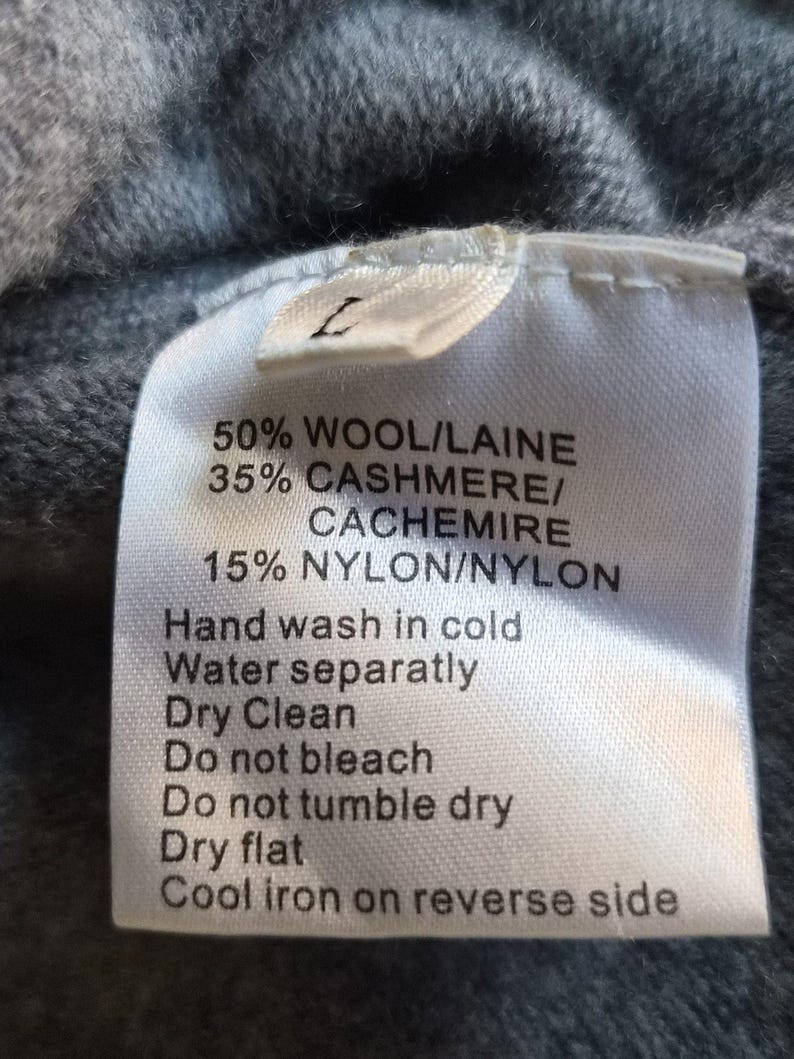 K&ouml;nnte beinhalten: Ein wei&szlig;es Textilpflegeetikett an einem grauen Strickstoff. Das Etikett zeigt die Gr&ouml;&szlig;e L und die Materialzusammensetzung von 50 % Wolle, 35 % Kaschmir und 15 % Nylon sowie spezifische Anweisungen zur Handw&auml;sche und Pflege des Kleidungsst&uuml;cks.