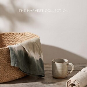 May include: A woven basket containing a folded cloth with a green and white tie-dye pattern, a ceramic mug with a speckled off-white finish, and a rolled beige fabric piece. The text THE HARVEST COLLECTION is visible at the top of the image.