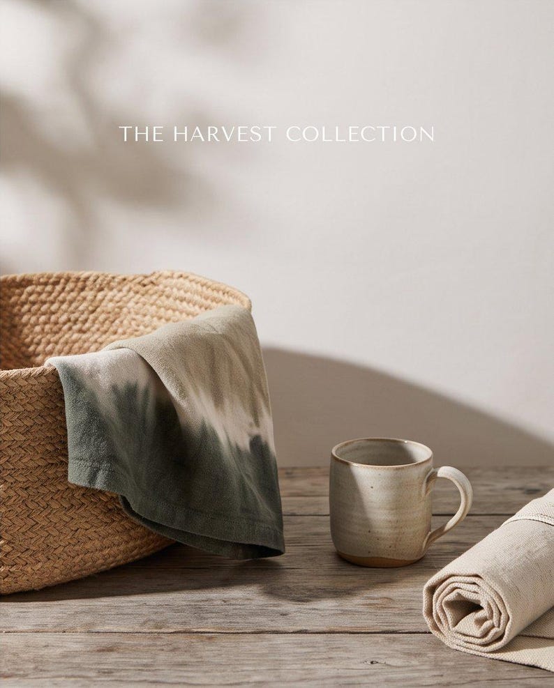 May include: A woven basket containing a folded cloth with a green and white tie-dye pattern, a ceramic mug with a speckled off-white finish, and a rolled beige fabric piece. The text THE HARVEST COLLECTION is visible at the top of the image.