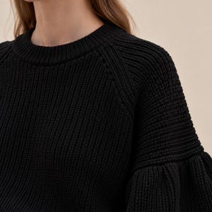 May include: A black long-sleeve sweater featuring a ribbed knit texture, a round neckline, and voluminous sleeves with a gathered cuff detail. The garment is paired with light blue trousers that have a visible waistband and belt loops.