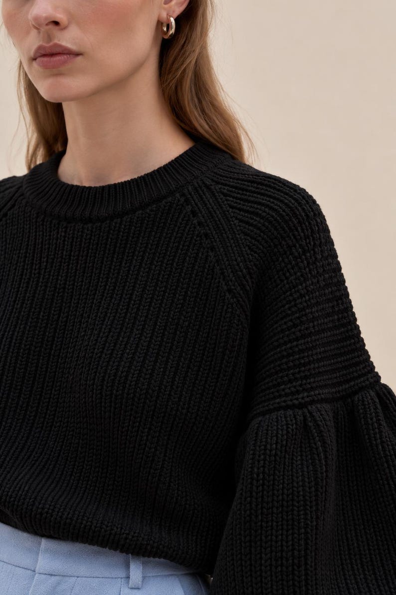 May include: A black long-sleeve sweater featuring a ribbed knit texture, a round neckline, and voluminous sleeves with a gathered cuff detail. The garment is paired with light blue trousers that have a visible waistband and belt loops.
