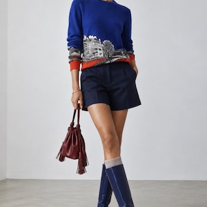 Puede incluir: Un su&eacute;ter azul real de manga larga con un estampado gr&aacute;fico arquitect&oacute;nico en blanco y negro y pu&ntilde;os naranjas, combinado con pantalones cortos azul marino, un peque&ntilde;o bolso de mano burdeos con borlas y botas altas de cuero azul marino.