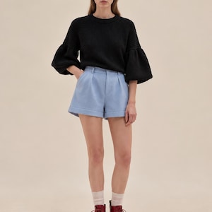 May include: A black ribbed knit sweater with voluminous sleeves paired with light blue tailored shorts featuring a front pleat and cuffed hems, worn with sheer white socks and dark red lace-up leather boots.