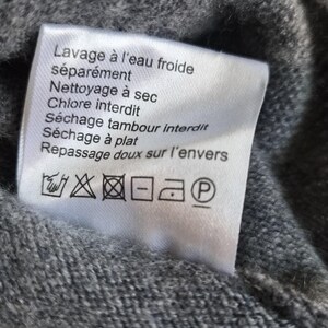 K&ouml;nnte beinhalten: Ein wei&szlig;es, rechteckiges Pflegeetikett, das an einem grauen Strickkleidungsst&uuml;ck befestigt ist. Das Etikett enth&auml;lt schwarzen Text auf Franz&ouml;sisch, darunter Lavage &agrave; l'eau froide s&eacute;par&eacute;ment, Nettoyage &agrave; sec, Chlore interdit, S&eacute;chage tambour interdit, S&eacute;chage &agrave; plat und Repassage doux sur l'envers.