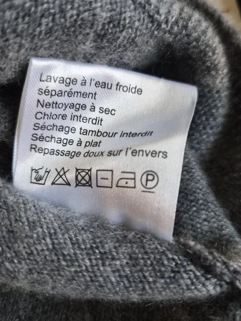 K&ouml;nnte beinhalten: Ein wei&szlig;es, rechteckiges Pflegeetikett, das an einem grauen Strickkleidungsst&uuml;ck befestigt ist. Das Etikett enth&auml;lt schwarzen Text auf Franz&ouml;sisch, darunter Lavage &agrave; l'eau froide s&eacute;par&eacute;ment, Nettoyage &agrave; sec, Chlore interdit, S&eacute;chage tambour interdit, S&eacute;chage &agrave; plat und Repassage doux sur l'envers.
