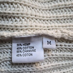 K&ouml;nnte beinhalten: Ein cremefarbener, grob gestrickter Stoff mit einem wei&szlig;en Etikett. Das Etikett zeigt den Text: 55% HEMP, 45% COTTON, 55% CHANVRE, 45% COTON, sowie ein separates, kleineres Etikett mit dem Buchstaben M f&uuml;r die Gr&ouml;&szlig;e Medium.