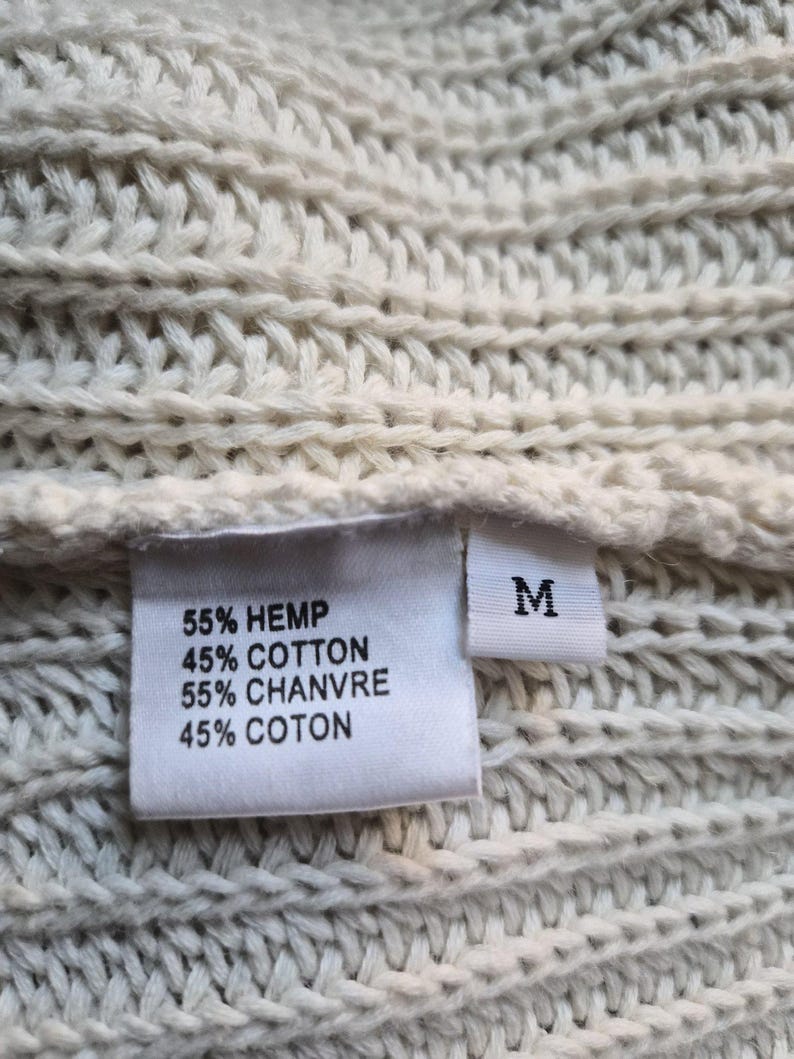 K&ouml;nnte beinhalten: Ein cremefarbener, grob gestrickter Stoff mit einem wei&szlig;en Etikett. Das Etikett zeigt den Text: 55% HEMP, 45% COTTON, 55% CHANVRE, 45% COTON, sowie ein separates, kleineres Etikett mit dem Buchstaben M f&uuml;r die Gr&ouml;&szlig;e Medium.