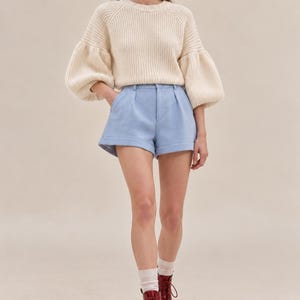 May include: A cream-colored ribbed knit sweater with voluminous long sleeves and a crew neckline, paired with light blue tailored shorts featuring a pleated front, cuffed hems, and a button closure, worn with white socks and dark red lace-up boots.