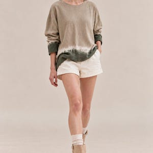 May include: A long-sleeve crewneck sweater in a light beige hue featuring a dark green tie-dye band at the hem and cuffs, paired with off-white cuffed shorts, light-colored ribbed socks, and beige suede ankle boots with side buckle closures.