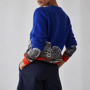 Puede incluir: Un su&eacute;ter de manga larga en color azul real con un estampado gr&aacute;fico en blanco y negro de un paisaje urbano en el dobladillo inferior, acabado con pu&ntilde;os de color naranja brillante. El su&eacute;ter se combina con pantalones cortos azul marino.