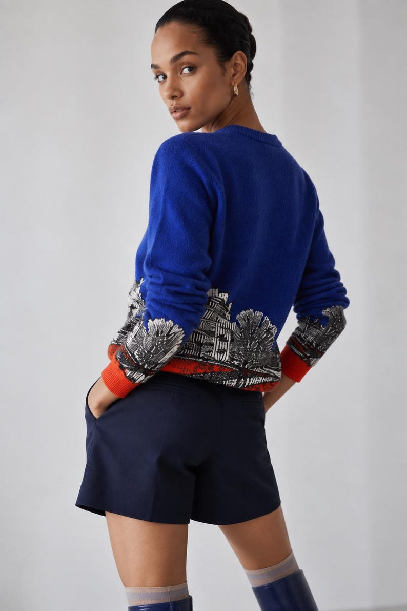 Puede incluir: Un su&eacute;ter de manga larga en color azul real con un estampado gr&aacute;fico en blanco y negro de un paisaje urbano en el dobladillo inferior, acabado con pu&ntilde;os de color naranja brillante. El su&eacute;ter se combina con pantalones cortos azul marino.